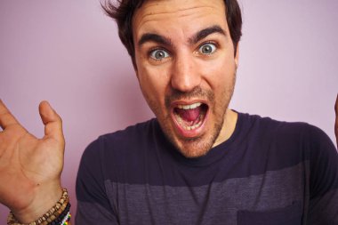 Yalnız pembe arka planda dikilen rahat tişörtlü genç yakışıklı adam mutlu ve heyecanlı, kazanan yüz ifadesi zaferi büyük bir gülümsemeyle kutluyor ve ellerini kaldırıyor.