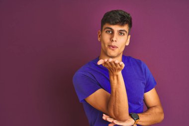 İzole edilmiş mor arka planda duran tişört giyen genç Hintli adam kameraya bakıp havada öpüşerek güzel ve seksi görünmeye çalışıyor. Aşk ifadesi.