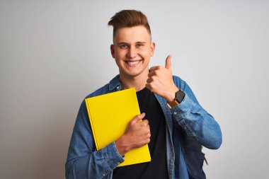 İzole edilmiş beyaz arka plan üzerinde kot gömlek giyen genç bir öğrenci. Gülümse ve işaret ver. Parmaklarınla başparmağını kaldır.
