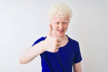 Mavi günlük tişörtler giyen genç albino bir adam izole edilmiş beyaz arka planda durup mutlu bir el hareketi yapıyor. Kameraya bakıp başarıyı gösteren ifade onaylanıyor.