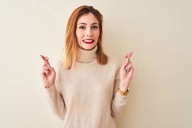 Kırmızı saçlı kadın, zarif balıkçı yaka kazak giyiyor. İzole edilmiş beyaz arka plan üzerinde duruyor. Umut ve gözler kapalı gülümseyerek el işareti yapıyor. Şans ve batıl inanç..