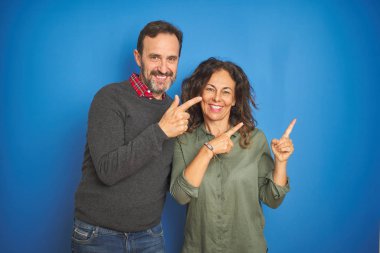 Güzel orta yaşlı çift, izole edilmiş mavi arka planda durup gülümsüyor ve kameraya bakıyor. İki eliyle ve parmaklarıyla kenarı işaret ediyor..
