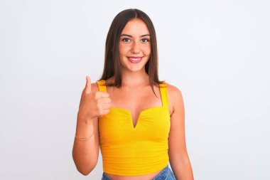 Sarı günlük tişört giyen genç ve güzel bir kız izole edilmiş beyaz arka planda durup mutlu bir el hareketi yapıyor. Kameraya bakıp başarıyı gösteren ifade onaylanıyor.