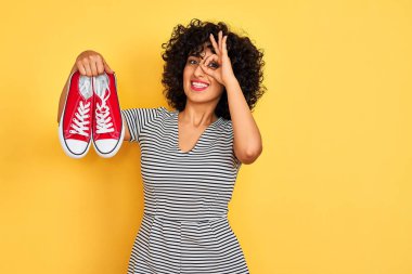 Kıvırcık saçlı genç bir Arap kadın izole edilmiş sarı arka planda spor ayakkabıları tutuyor, gülümsüyor ve el pençe divan duruyor.
