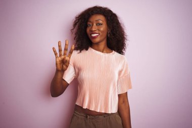 Genç Afro-Amerikan kadın tişört giyiyor pembe arka plan üzerinde duruyor ve parmaklarını dört numarayı gösterirken kendinden emin ve mutlu bir şekilde gülümsüyor..