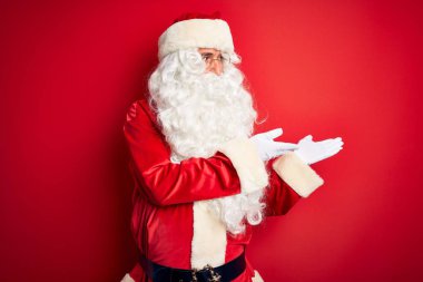 Orta yaşlı, Noel Baba kostümü giymiş yakışıklı bir adam izole edilmiş kırmızı arka planda duruyor. Elinde avuç içi açık, fotokopi alanı gösteren, mutlu bir gülümseme reklamı sunuyor.