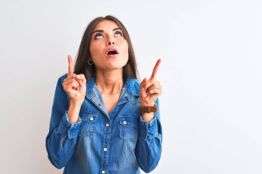 İzole edilmiş beyaz arka plan üzerinde duran sıradan bir kot gömlek giyen genç ve güzel bir kadın şaşırmış ve şaşırmış bir şekilde yukarı bakıyor ve parmaklarıyla işaret ediyor ve kollarını kaldırıyor..