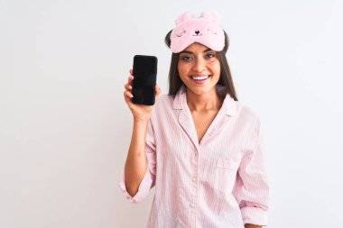 Uyku maskesi takan genç bir kadın, elinde akıllı telefon, izole edilmiş beyaz bir arka plan, mutlu bir yüz, kendine güvenen bir gülümseme ve dişlerini gösteren bir gülümsemeyle.