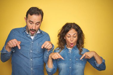 Güzel orta yaşlı çift, izole edilmiş sarı arka planda dikiliyorlar. İlan veren parmaklarıyla işaret ediyorlar. Şaşırmış bir yüz ve açık bir ağız.