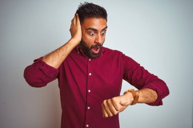 Kırmızı şık gömlekli genç Hintli adam, gri arka planda dikiliyor. Endişeli saate bakıyor, geç kalmaktan korkuyor.