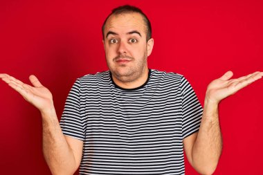Çizgili donanma tişörtü giyen genç adam izole edilmiş kırmızı arka plan üzerinde duruyor. Kolları ve elleri havada, kafası karışmış bir ifade takınıyor. Şüphe kavramı.