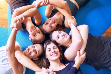 Yoga yapan genç ve güzel bir kadın grubu yerde daireler çiziyor. Spor salonunda el ele tutuşurken mutlu bir şekilde gülümsüyor.