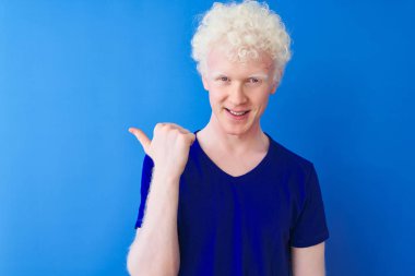 Genç, albino, günlük tişört giyen, mavi arka planda duran mutlu yüzüyle gülümseyen ve baş parmağıyla yan tarafı gösteren sarışın bir adam..