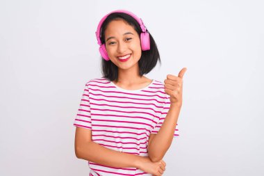 Güzel Çinli kadın müzik dinliyor, kulaklık takıyor, beyaz arka planda mutlu, gülümsüyor, işaret veriyor, parmaklarıyla başparmağını kaldırıyor, mükemmel bir işaret.