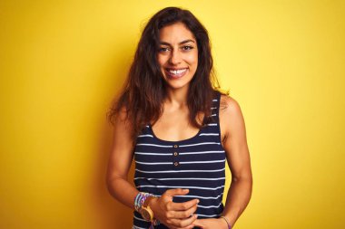İzole edilmiş sarı arka plan üzerinde duran çizgili tişört giyen genç ve güzel bir kadın ellerini kavuşturup gülümsüyor. Başarı ve iyimser