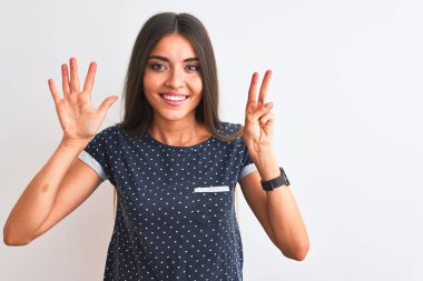 İzole edilmiş beyaz arka plan üzerinde duran mavi günlük tişört giyen genç ve güzel bir kadın yedinci parmaklarını gösterirken kendinden emin ve mutlu bir şekilde gülümsüyor..