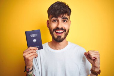 Alman pasaportu takan dövmeli genç adam sarı arka planda gururla bağırıyor ve zaferi ve başarıyı kutluyor.