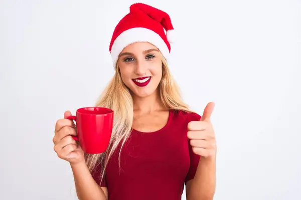 漂亮的女人戴着圣诞圣诞礼帽 在与世隔绝的白色背景上喝咖啡 开心地笑着做着好的手势 用手指竖起大拇指 很好的标志 — 图库照片