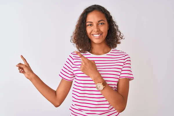 年轻的巴西女人穿着粉色条纹T恤 站在孤立的白色背景上微笑着 看着摄像机 用双手和手指指向旁边 — 图库照片