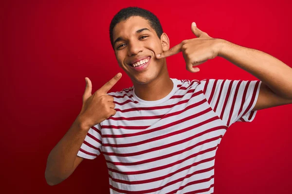 年轻英俊的阿拉伯男子穿着条纹T恤 披着孤立的红色背景 笑容满面 用手指 牙齿和嘴指尖 牙齿健康概念 — 图库照片