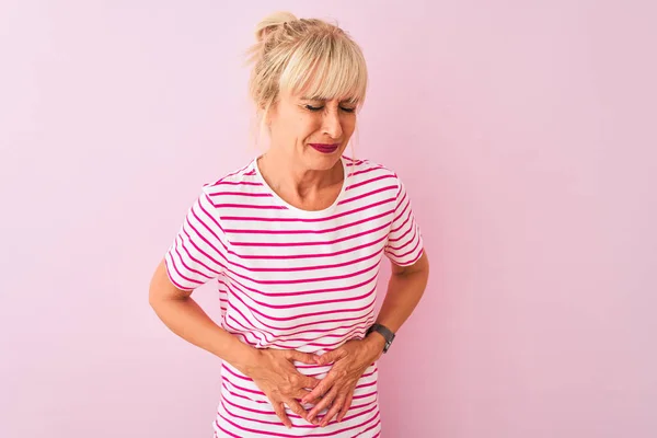 Middle age woman wearing striped t-shirt standing over isolated pink background with hand on stomach because indigestion, painful illness feeling unwell. Ache concept.