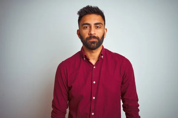 Young indian man wearing red elegant shirt standing over isolated grey background Relaxed with serious expression on face. Simple and natural looking at the camera.