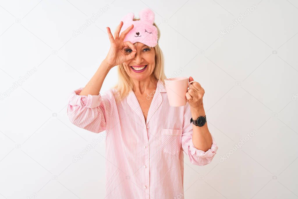 Middle age woman wearing sleep mask pajama drinking coffee over isolated white background with happy face smiling doing ok sign with hand on eye looking through fingers
