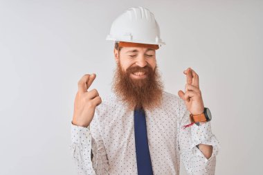 İzole edilmiş beyaz arka plan üzerinde güvenlik kaskı takan genç kızıl saçlı mimar umut ve gözlerin kapalı bir şekilde gülümseyerek işaret ediyor. Şans ve batıl inanç..