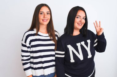 İzole edilmiş beyaz arka planda dikilen gündelik kıyafetler giyen genç ve güzel kadınlar üç numaralı parmaklarıyla işaret ederken kendinden emin ve mutlu bir şekilde gülümsüyorlar..