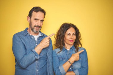 Güzel bir orta yaşlı çift, izole edilmiş sarı arka planda dikiliyorlar. Elleriyle işaret ediyorlar. Reklam yapıyorlar, ciddi ve sakin bir yüz.