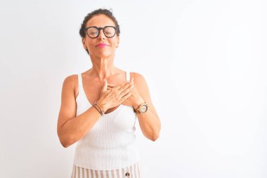 Sıradan tişört ve gözlük takan orta yaşlı bir kadın izole edilmiş beyaz arka planda duruyor elleri göğsünde, gözleri kapalı ve yüzünde minnet dolu bir jest ile gülümsüyor. Sağlık kavramı.