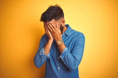 İzole edilmiş sarı arka plan üzerinde duran kot gömlek giyen genç Hintli bir adam. Ağlarken yüzünü ellerle kapatan üzgün bir ifade. Depresyon kavramı.