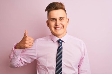 Genç yakışıklı iş adamı gömleği ve kravatıyla soyutlanmış pembe arka planda dikiliyor. Gururlu görünüyor, gülümsüyor, baş parmak işareti yapıyor.