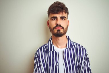 İzole edilmiş beyaz arka plan üzerinde duran çizgili gömlekli genç adam şüpheci ve gergin, yüzünde çapraz kollu tasvip etmeyen bir ifade var. Negatif kişi.