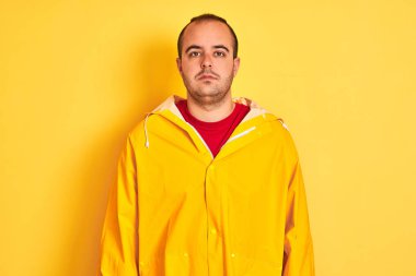 İzole edilmiş sarı arka planda duran yağmurluk giyen genç adam yüzünde ciddi bir ifadeyle rahatlamış. Basit ve doğal bir şekilde kameraya bakıyorsun..