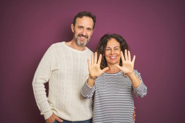 Güzel bir orta yaşlı çift, mor arka plan üzerine kışlık süveter giyip parmaklarını on numara gösterirken mutlu ve kendinden emin bir şekilde gülümsüyor..