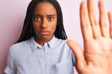 Genç Afro-Amerikan kadın çizgili gömlek giyiyor pembe arka planda duruyor açık elle dur işareti yapıyor ciddi ve kendinden emin bir ifadeyle, savunma jesti