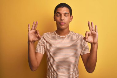 İzole edilmiş sarı arka planda duran çizgili tişört giyen yakışıklı Arap adam rahatla ve gözleri kapalı meditasyon hareketi yaparken parmaklarınla gülümse. Yoga konsepti.