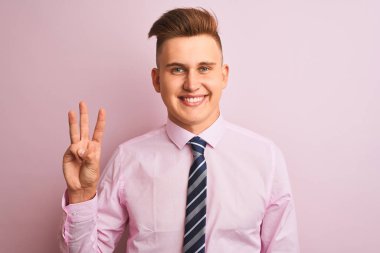 Genç yakışıklı iş adamı gömleği ve kravatıyla pembe arka planda dikiliyor ve üç numaralı parmaklarıyla işaret ederken kendinden emin ve mutlu bir şekilde gülümsüyor..