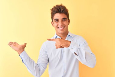 Zarif bir gömlek giyen genç yakışıklı iş adamı, izole edilmiş sarı arka planda hayret ediyor ve kameraya gülümsüyor, eliyle ve parmağıyla işaret ediyor..