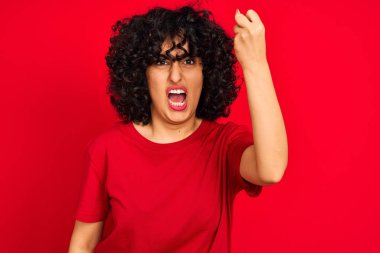 Kıvırcık saçlı genç Arap kadın izole edilmiş kırmızı arka plan üzerine gündelik tişört giyiyor kızgın ve öfkeyle bağırırken yumruğunu kaldırıyor. Öfke ve agresif kavram.