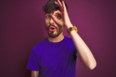 İzole edilmiş mor arka plan üzerinde duran dövmeli genç adam şaşırmış yüz ve parmaklarından bakan gözle şok edici hareketler yapıyor. İnançsız ifade.