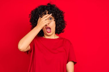 Kıvırcık saçlı genç bir Arap kadın izole edilmiş kırmızı arka plan üzerinde günlük tişört giyiyor. Şok olmuş yüz ve gözlerle, utanmış bir ifadeyle parmaklarının arasından bakıyor..