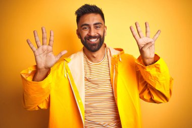 İzole edilmiş sarı arka planda duran yağmurluk giyen genç Hintli adam dokuz numaralı parmaklarıyla işaret ederken kendinden emin ve mutlu gülümsüyor..