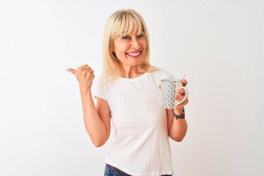 Orta yaşlı bir kadın izole edilmiş beyaz arka planda dikilmiş bir fincan kahve içiyor ve gülümseyen bir gülümsemeyle baş parmağıyla onu işaret ediyor.