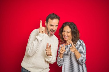 İzole edilmiş kırmızı arka plan üzerine kışlık süveter giyen güzel orta yaşlı çift mutlu ve komik yüzlü parmaklarını kameraya doğrultuyor. İyi enerji ve titreşimler.
