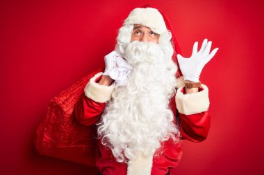 Noel Baba kostümü giymiş orta yaşlı bir adam kırmızı arka planda hediyelerle dolu bir çanta taşıyor. Mutlu ve heyecanlı. Kazanan ifade zaferi kutluyor.