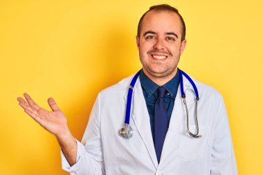 Ceket ve stetoskop giyen genç doktor izole edilmiş sarı arka planda dikiliyor neşeli bir şekilde gülümsüyor ve avuç içi kameraya bakıyor..