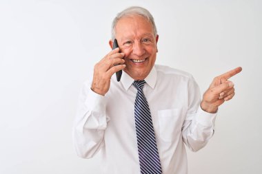 Üst düzey gri saçlı işadamı, izole edilmiş beyaz arka planda akıllı telefondan konuşuyor. Elleri ve parmakları kenarı işaret ederek çok mutlu.