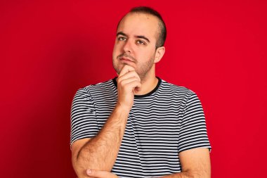 Çizgili donanma tişörtü giyen genç bir adam izole edilmiş kırmızı arka plan üzerinde duruyor ve elleri çenesinde soru sormayı düşünüyor, düşünceli bir ifade. Düşünceli bir yüzle gülümsüyor. Şüphe kavramı.